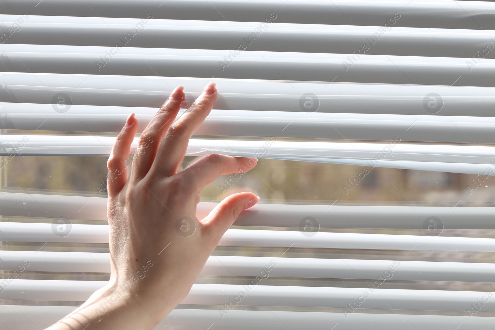 Photo of Woman separating slats of white blinds indoors, closeup