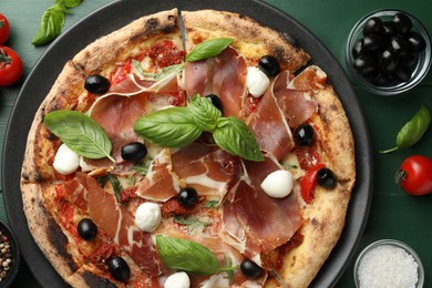 Tasty pizza with cured ham, olives, mozzarella cheese, tomatoes and arugula on green wooden table, flat lay