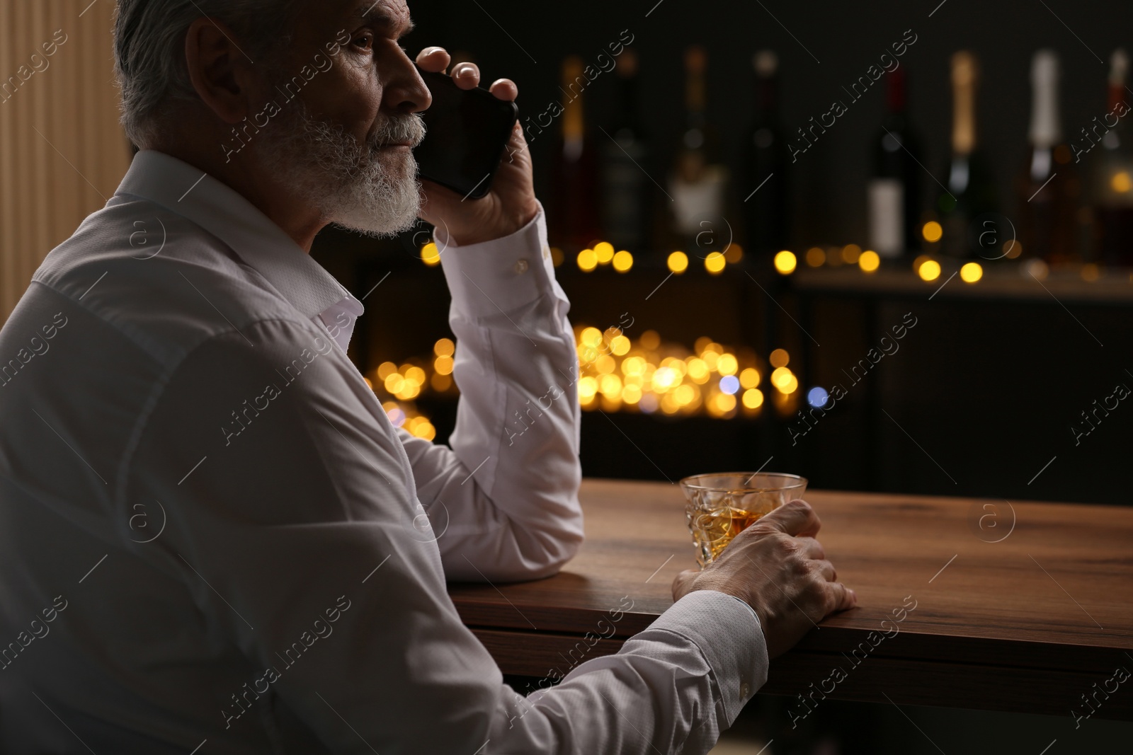 Photo of Senior man with glass of whiskey talking on phone at bar counter against blurred lights. Space for text