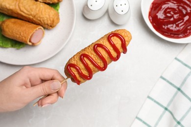 Photo of Woman holding delicious corn dog with ketchup at light table, closeup
