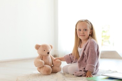Pretty little girl with book and teddy bear on floor in room