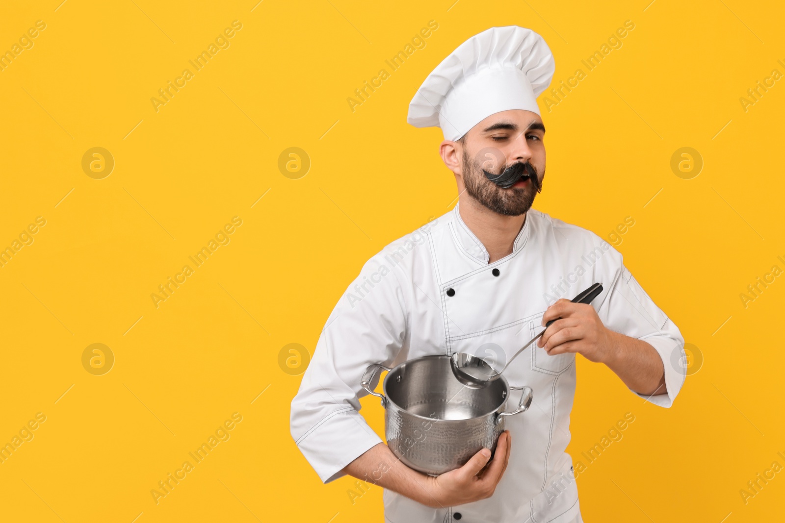 Photo of Professional chef with funny artificial moustache holding cooking pot and ladle on yellow background. Space for text