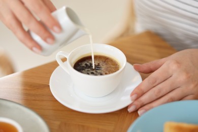 Photo of Breakfast. Woman pouring cream into cup of coffee at wooden table, closeup