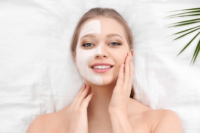 Beautiful woman with clay facial mask and tropical leaf on white fabric, above view