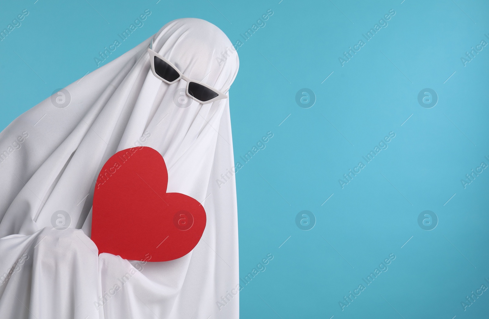Photo of Cute ghost. Person covered with white sheet in sunglasses holding red heart on light blue background, space for text
