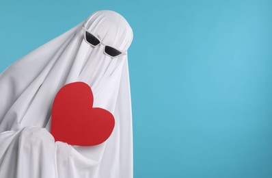 Cute ghost. Person covered with white sheet in sunglasses holding red heart on light blue background, space for text