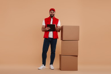 Happy young courier with clipboard and stack of parcels on light brown background