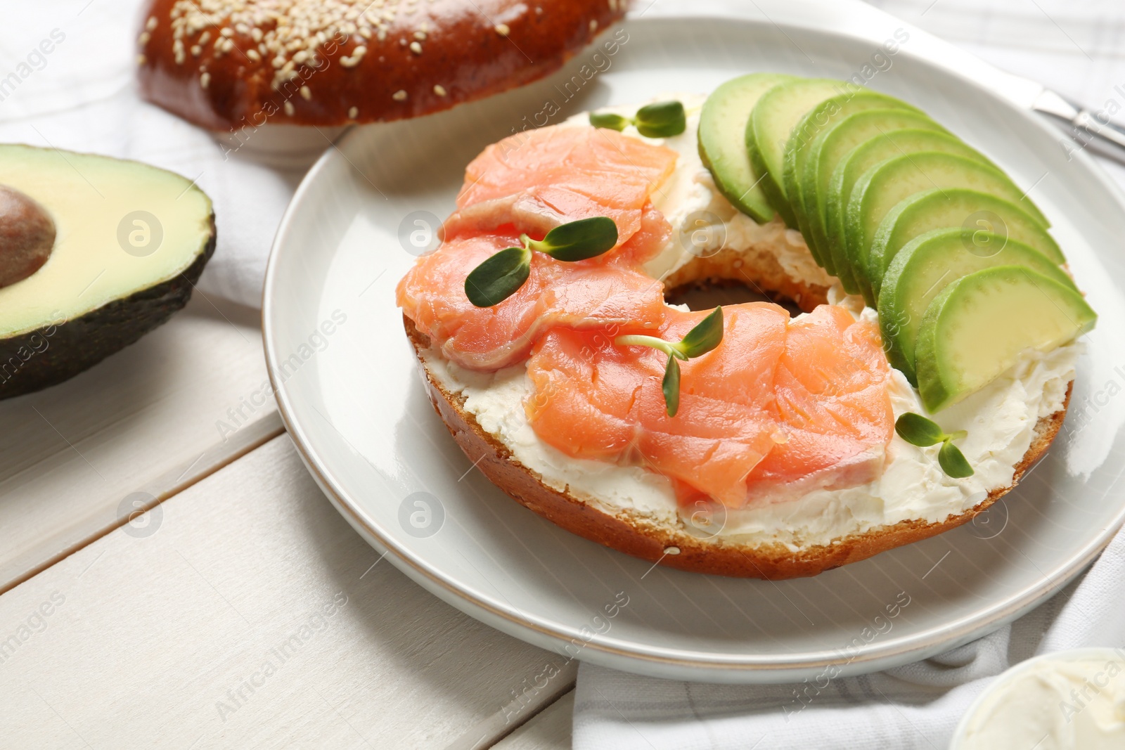 Photo of Delicious bagel with cream cheese, salmon and avocado on white wooden table, closeup