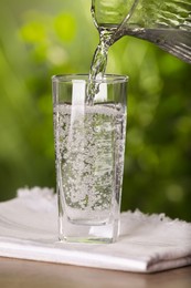 Pouring water from jug into glass on wooden table outdoors, closeup