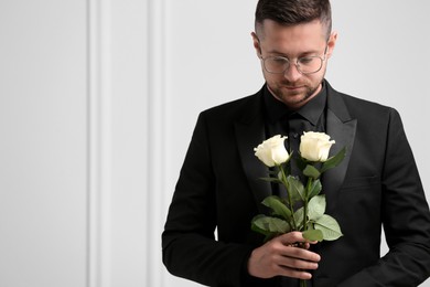 Sad man with rose flowers near white wall, space for text. Funeral ceremony