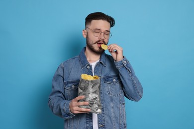 Handsome young man eating tasty potato chips on light blue background