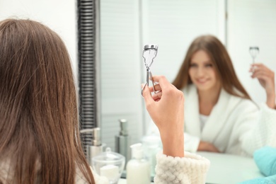 Photo of Young woman with eyelash curler near mirror in bathroom, closeup