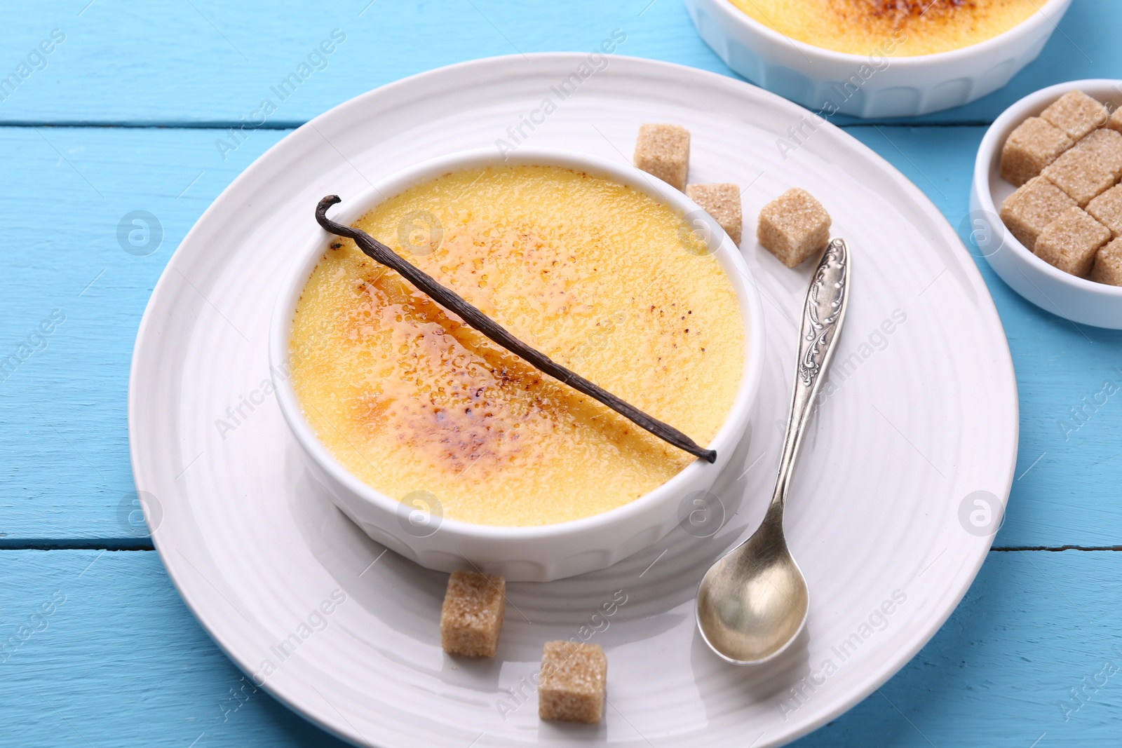 Photo of Delicious creme brulee in bowl, vanilla pod, sugar cubes and spoon on light blue wooden table