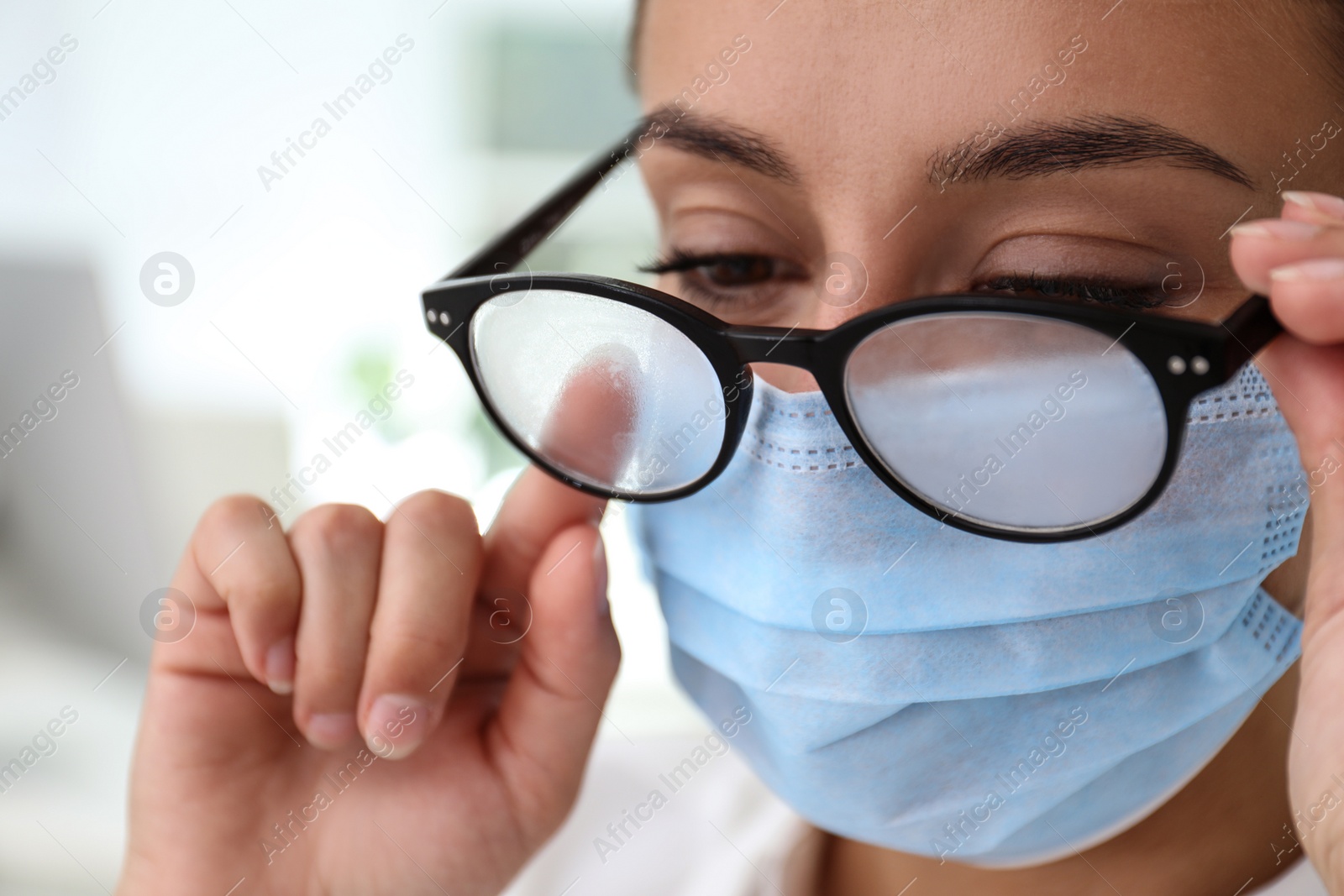 Photo of Woman wiping foggy glasses caused by wearing medical mask indoors, closeup