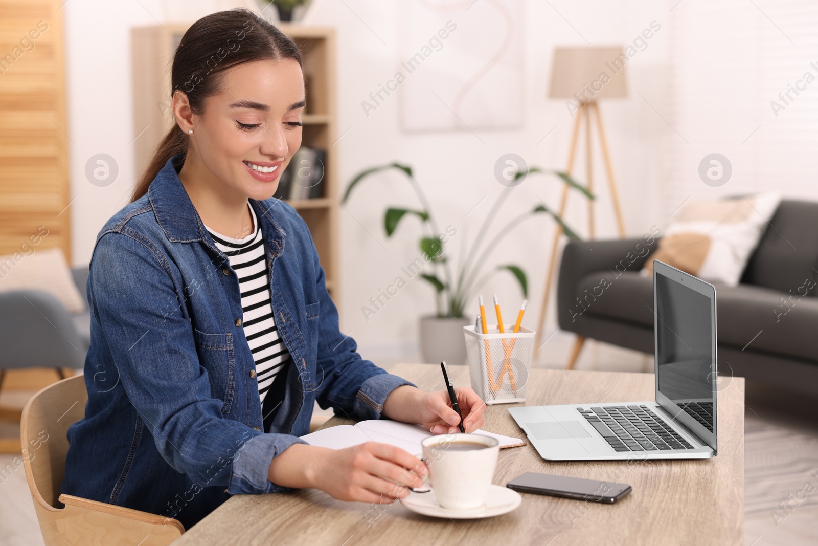 Photo of Young woman with cup of drink writing in notebook at wooden table indoors