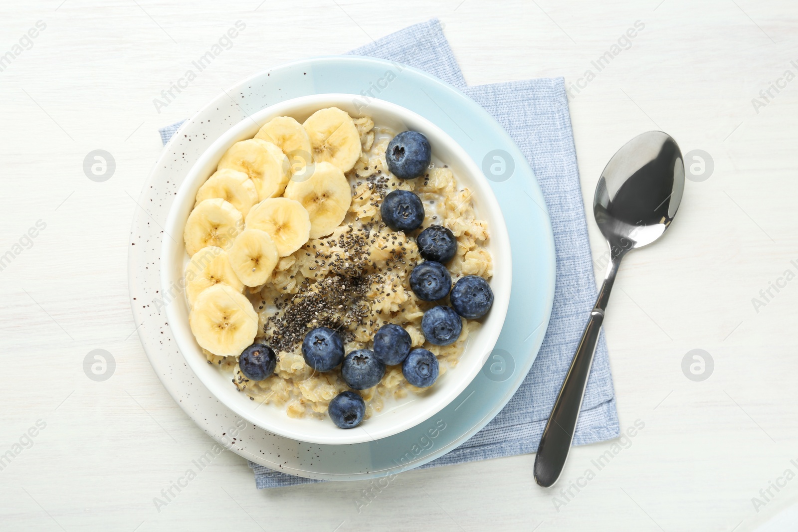 Photo of Tasty oatmeal with banana, blueberries and chia seeds served in bowl on white wooden table, top view