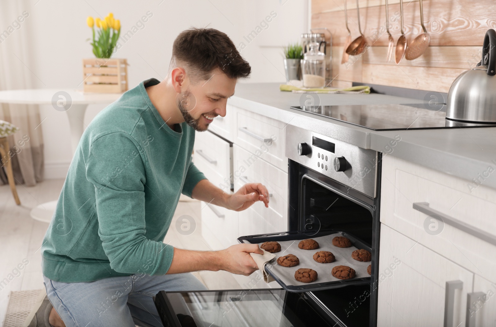 Photo of Handsome man taking out tray of baked cookies from oven in kitchen