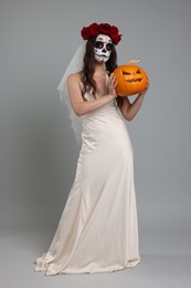 Photo of Young woman in scary bride costume with sugar skull makeup, flower crown and carved pumpkin on light grey background. Halloween celebration