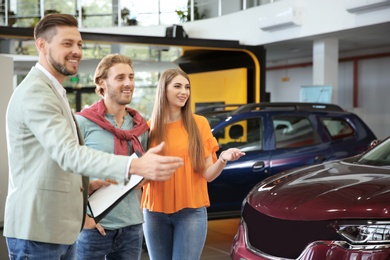 Salesman with clipboard consulting young couple in modern car dealership