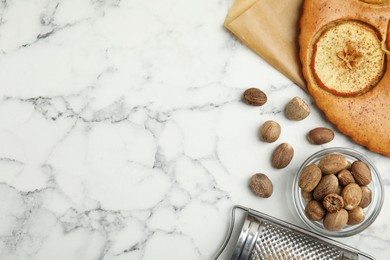 Nutmeg seeds, grater and tasty apple pie on white marble table, flat lay. Space for text