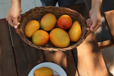 Woman with wicker basket of tasty mangoes at wooden table, top view