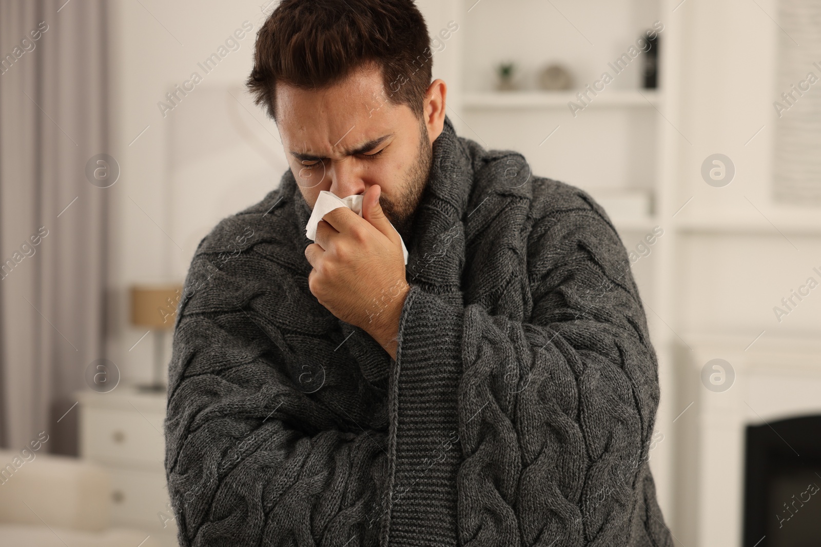 Photo of Sick man wrapped in blanket with tissue blowing nose at home. Cold symptoms