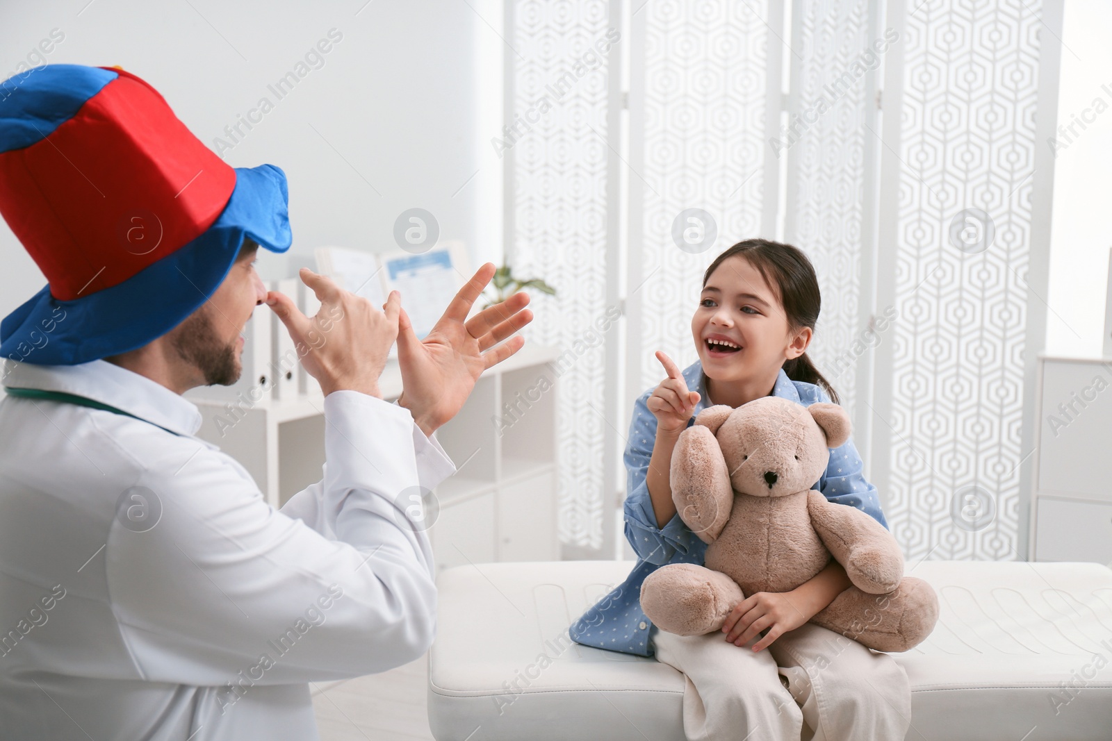 Photo of Pediatrician in funny hat playing with little girl during visit at hospital