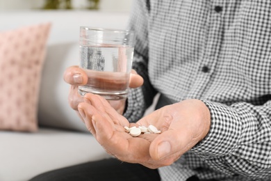 Senior man holding pills and glass of water indoors, closeup