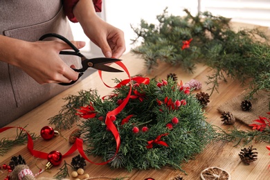 Photo of Florist making beautiful Christmas wreath with berries and red ribbon at wooden table indoors, closeup