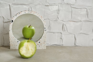Photo of Bitten green apple near mirror with reflection of whole fruit on table, space for text