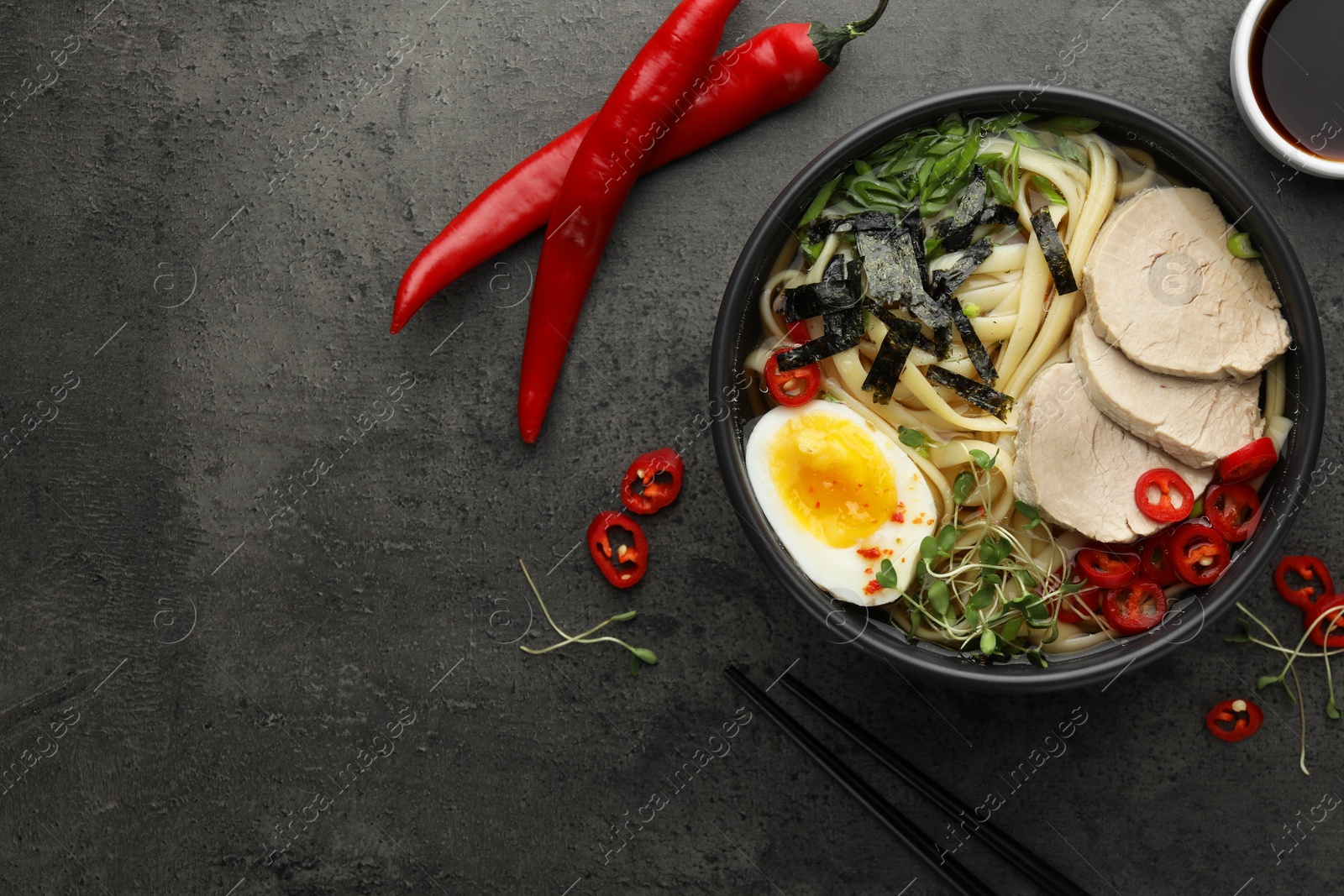 Photo of Delicious ramen with meat in bowl and ingredients on grey textured table, flat lay. Space for text. Noodle soup