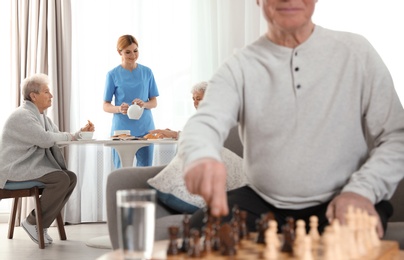 Nurse serving breakfast to elderly women and aged man playing chess at retirement home. Assisting senior people