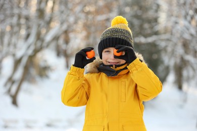 Cute little boy covering eye with tangerine in snowy park on winter day