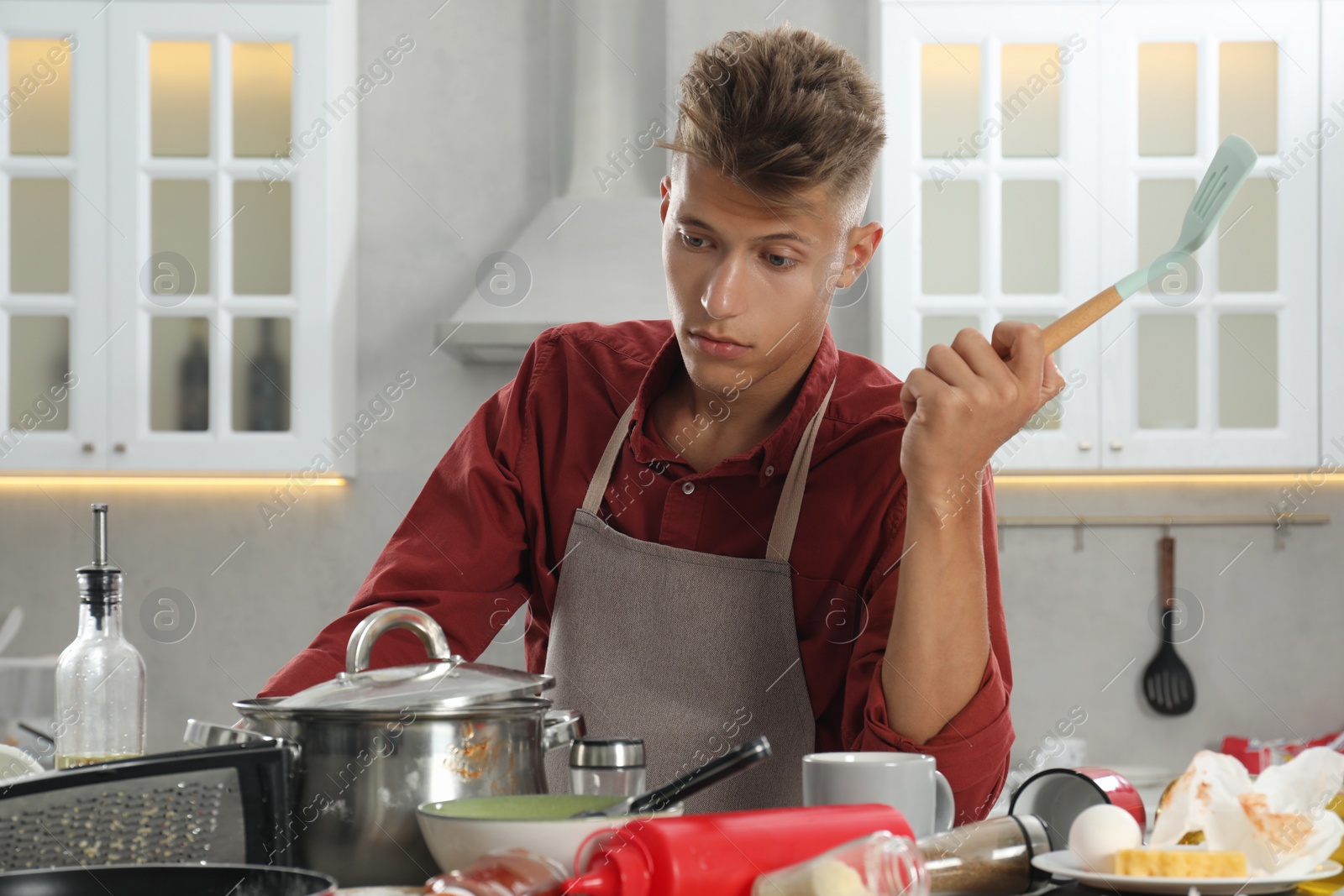 Photo of Upset man with spatula in messy kitchen. Many dirty dishware, utensils and food leftovers on table