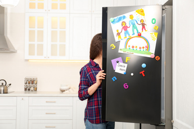 Photo of Woman opening refrigerator door with child's drawings, notes and magnets in kitchen. Space for text
