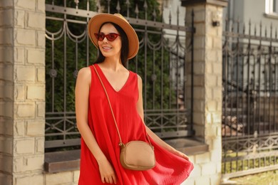 Beautiful young woman in stylish red dress and sunglasses walking on city street, space for text