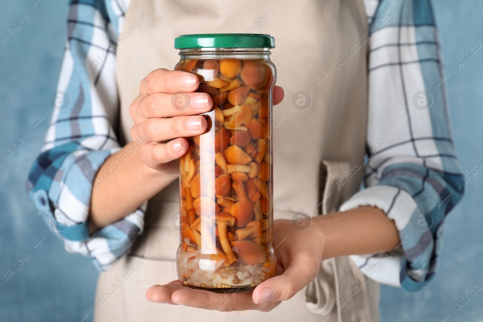 Photo of Woman holding jar with pickled mushrooms against blue background, closeup