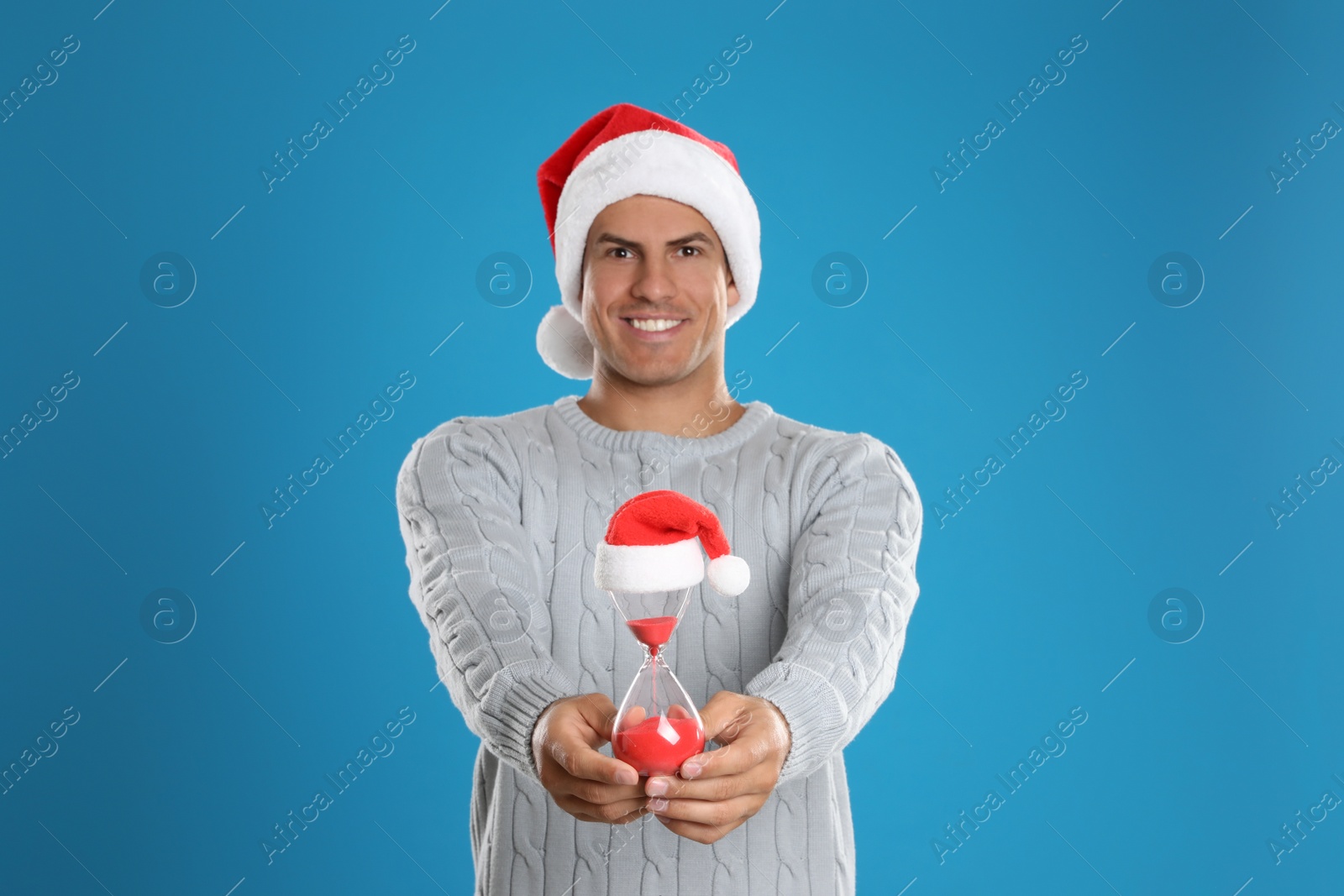 Photo of Man in Santa hat with hourglass on blue background. Christmas countdown