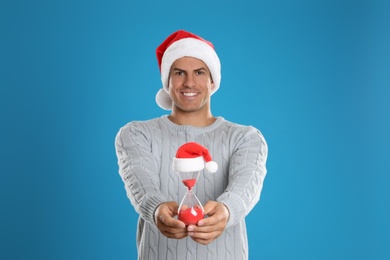 Photo of Man in Santa hat with hourglass on blue background. Christmas countdown