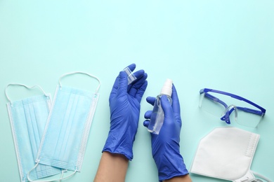 Person in gloves with hand sanitizer surrounded by medical items on light blue background, top view