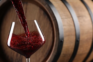 Pouring red wine into glass near wooden barrel, closeup. Space for text