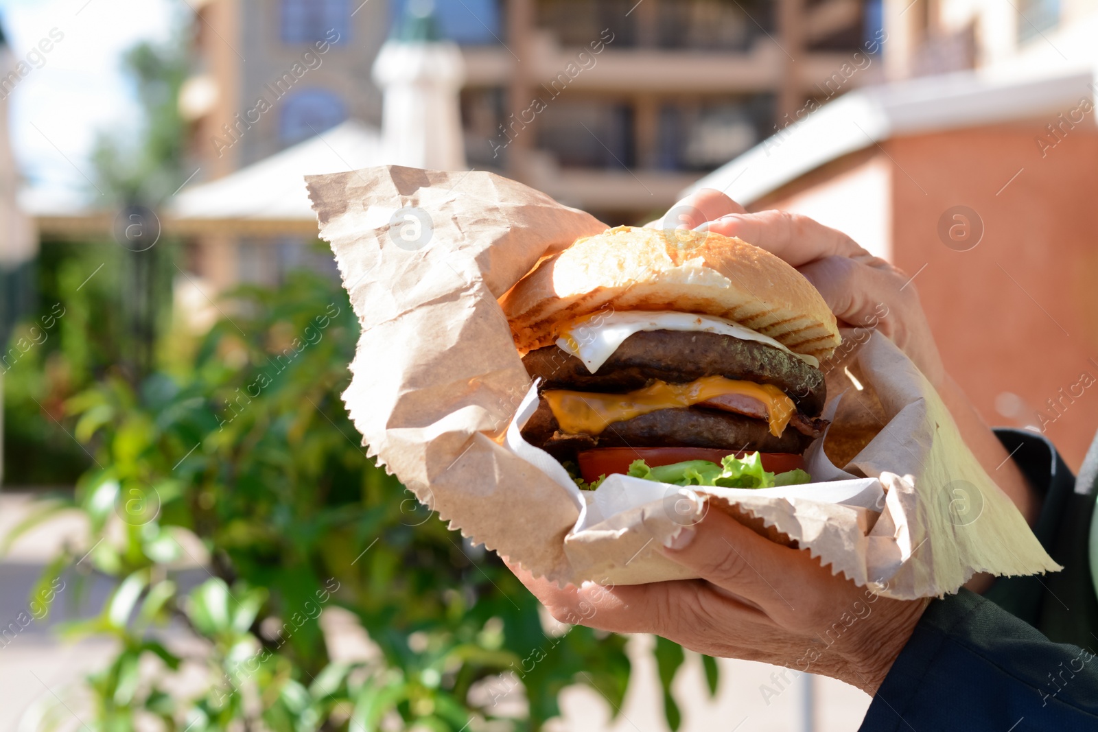 Photo of Woman holding delicious burger in paper wrap on city street, closeup