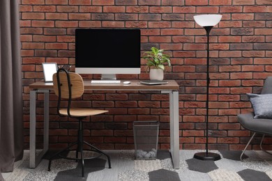 Photo of Cosy workspace with computer on desk, chair and floor lamp near brick wall at home