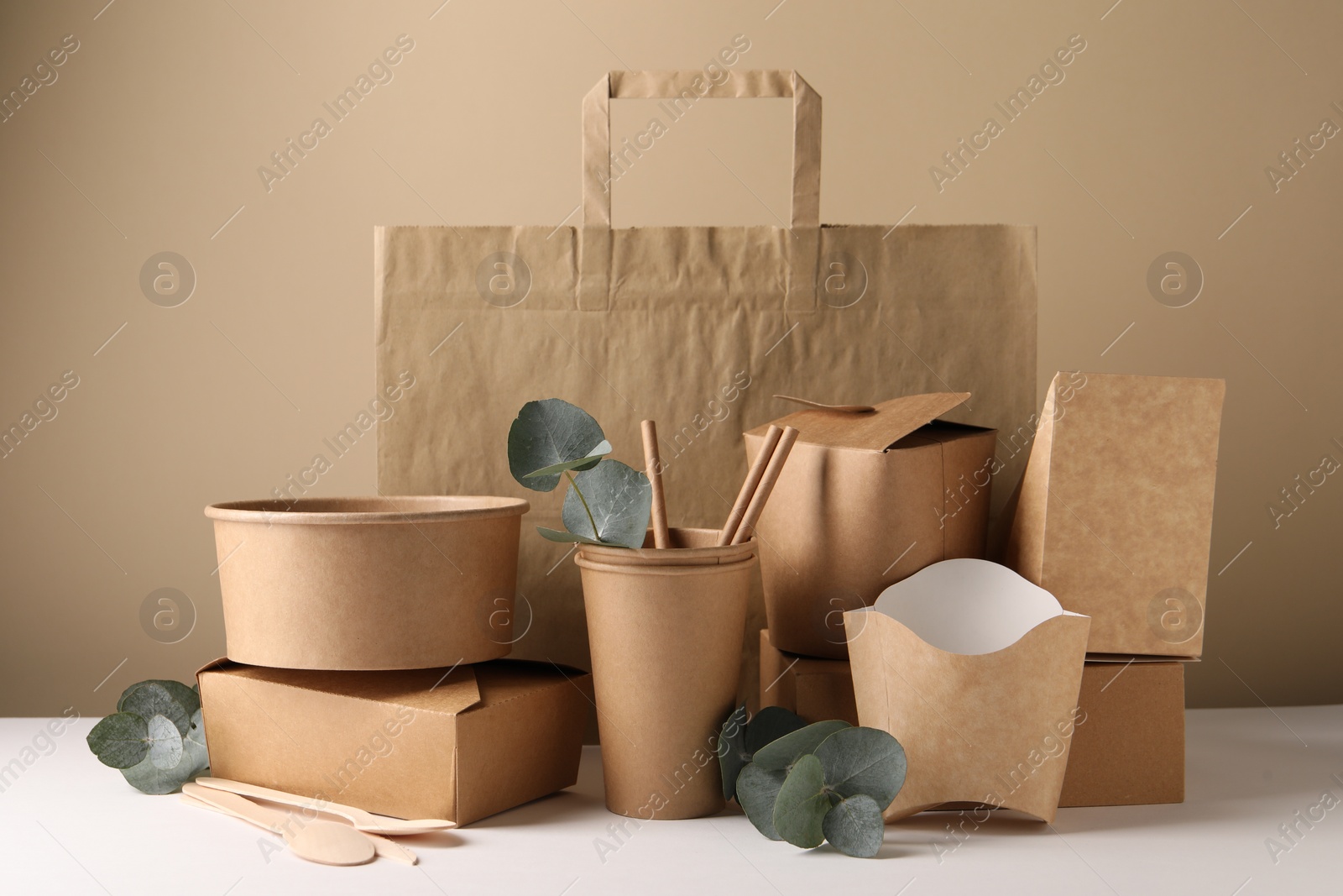 Photo of Eco friendly food packaging. Paper containers, tableware, bag and eucalyptus branches on white table against beige background