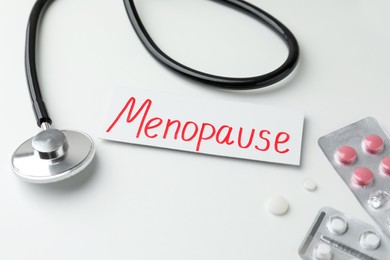 Card with word Menopause, pills and stethoscope on white table, closeup