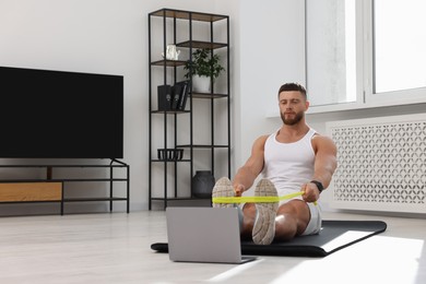 Photo of Muscular man doing exercise with elastic resistance band near laptop on mat at home