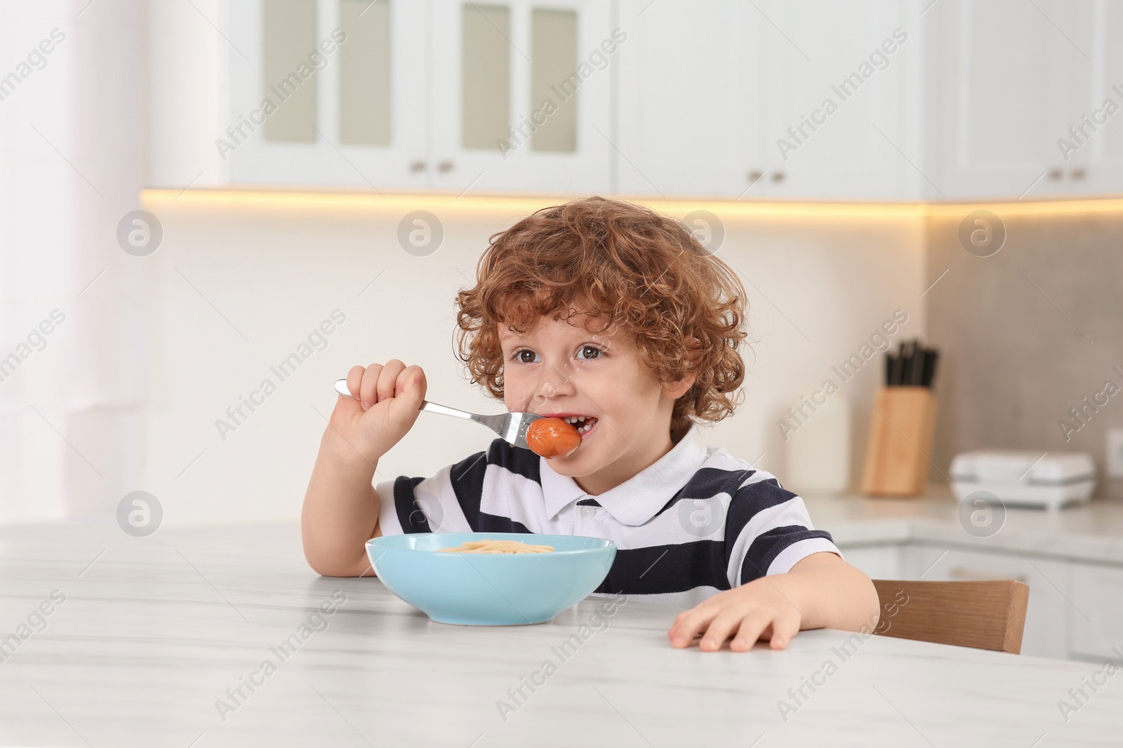 Photo of Cute little boy eating sausage and pasta at table in kitchen