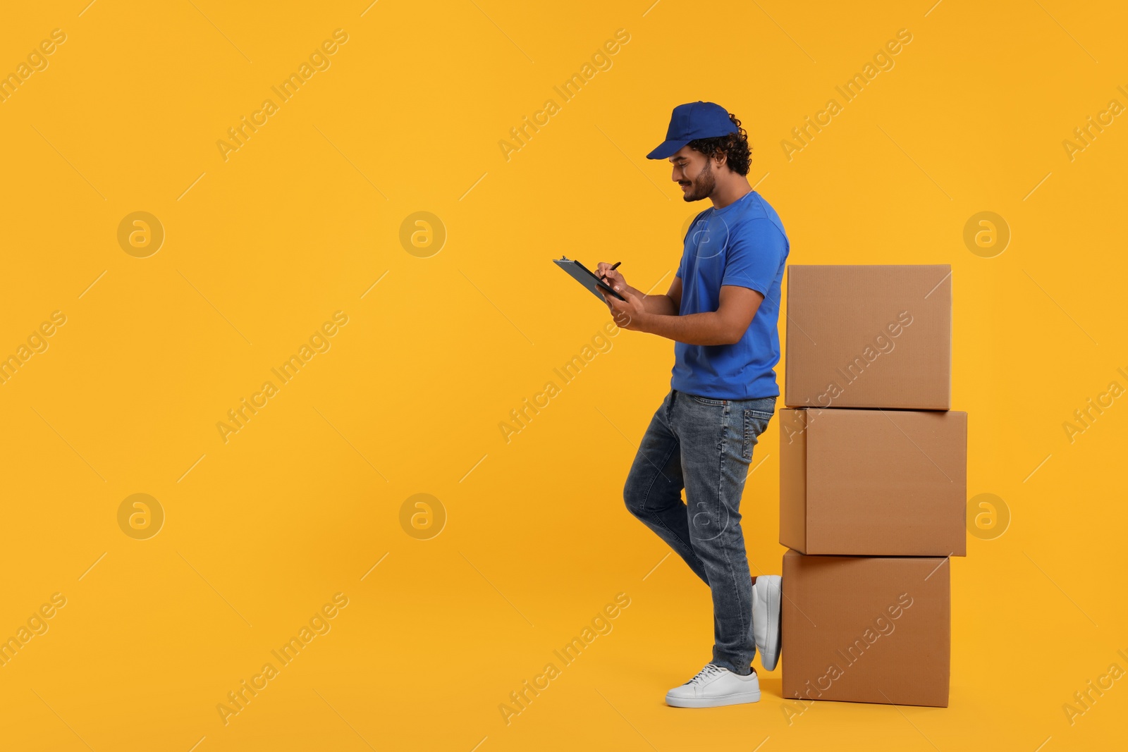 Photo of Happy courier with clipboard and stack of parcels on orange background, space for text