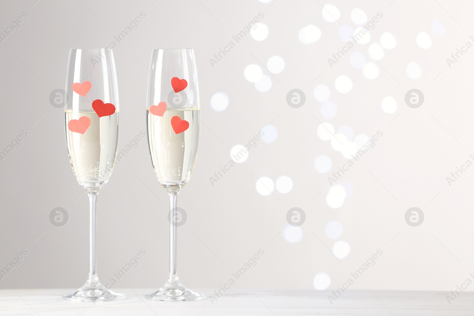 Photo of Glasses of champagne with paper hearts on white table against blurred lights. Space for text