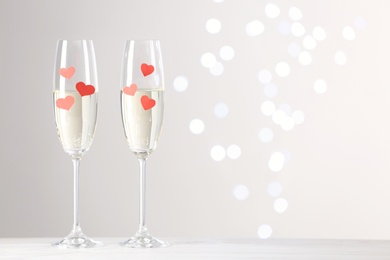 Photo of Glasses of champagne with paper hearts on white table against blurred lights. Space for text